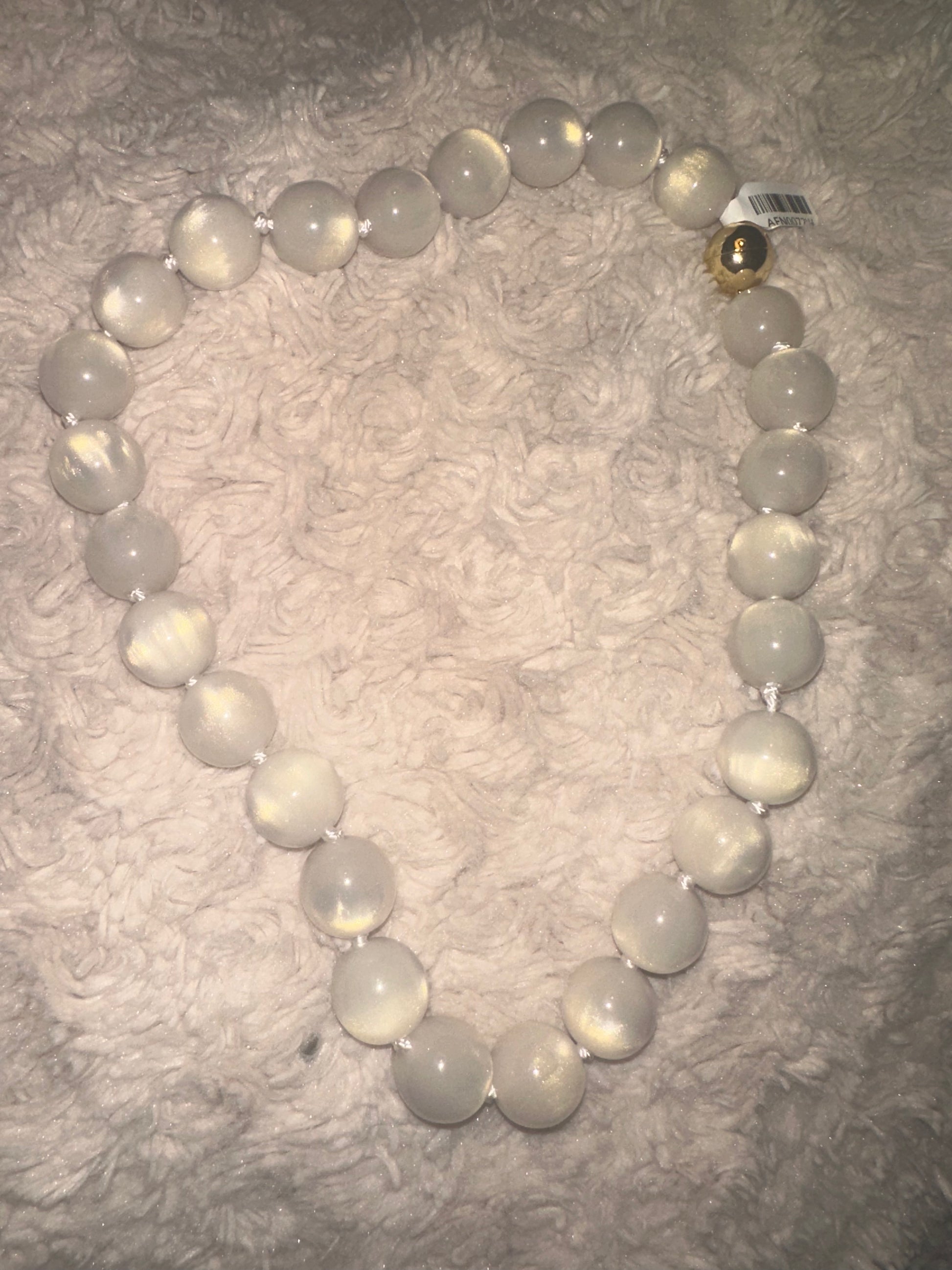 A beaded necklace made of large, smooth, milky white oval beads with a small metallic gold accent bead, displayed on a textured, light beige faux fur surface.