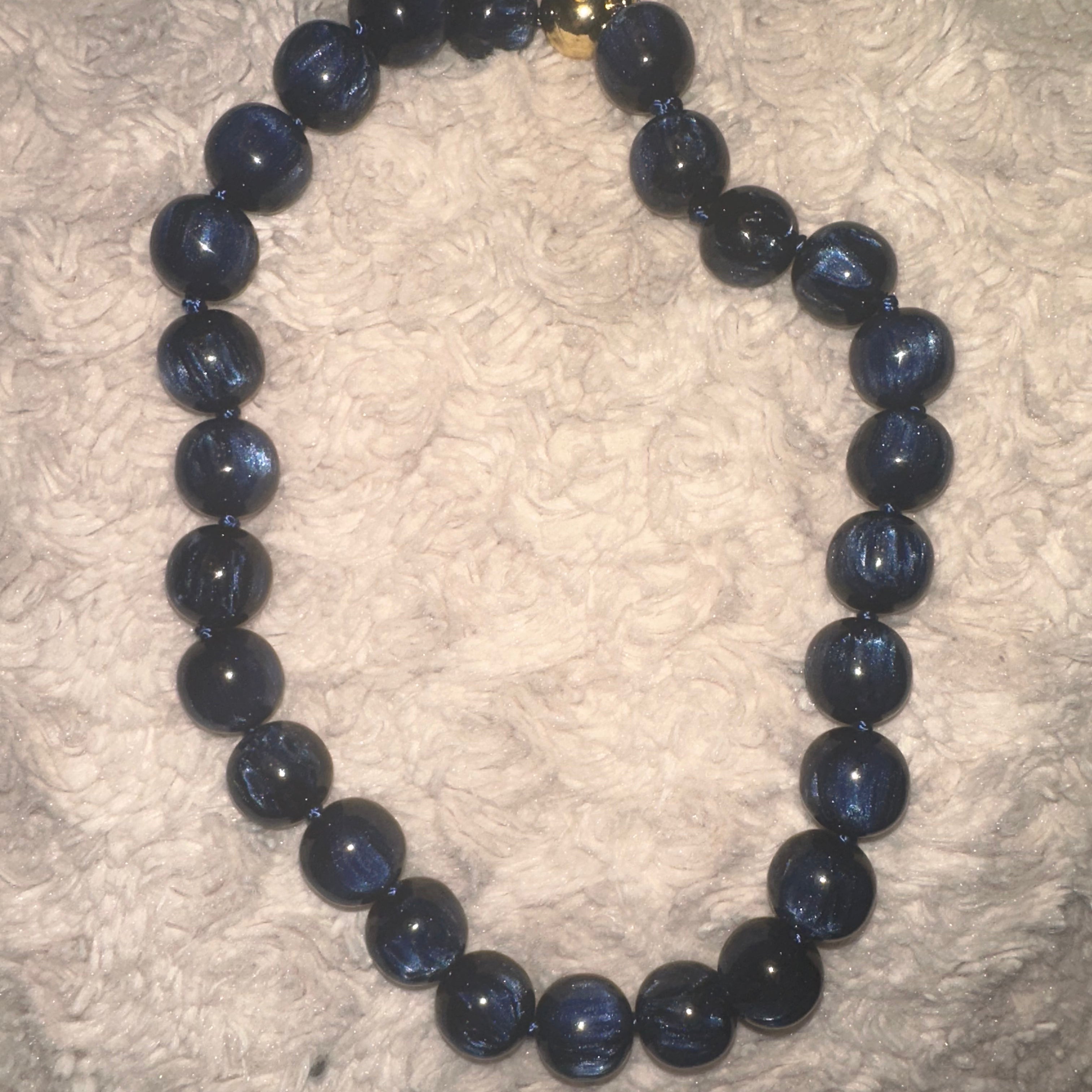 A beaded necklace made of dark blue, semi-translucent round beads with a small gold accent bead near the clasp, displayed on a textured white fabric background.