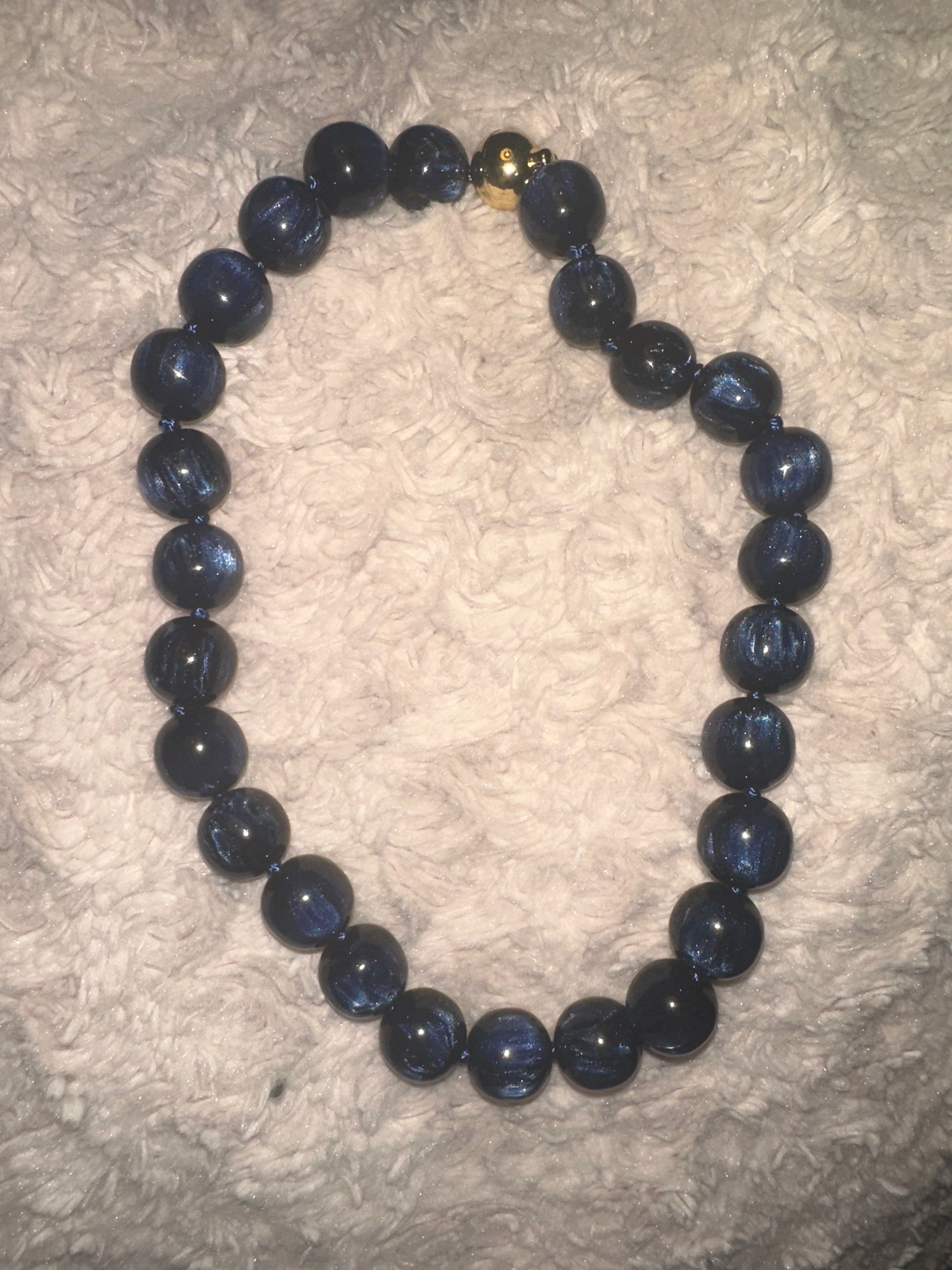 A beaded necklace made of dark blue, semi-translucent round beads with a small gold accent bead near the clasp, displayed on a textured white fabric background.