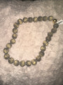 A beaded necklace made of large, round, metallic grey-toned beads with a swirled, tiger-eye-like pattern, displayed on a textured gray plush blanket. A small barcode tag is attached near the top right.
