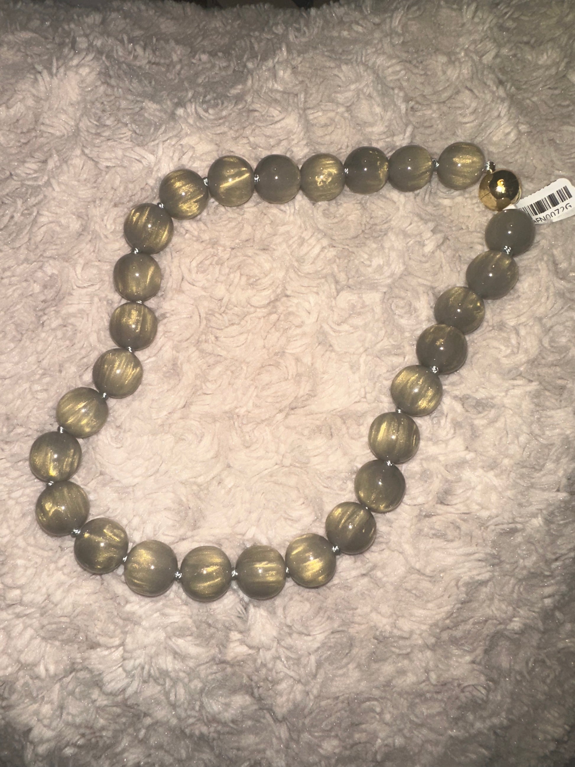 A beaded necklace made of large, round, metallic grey-toned beads with a swirled, tiger-eye-like pattern, displayed on a textured gray plush blanket. A small barcode tag is attached near the top right.