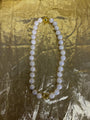 A beaded necklace made of round white pearl-like beads with iridescent blue flashes, accented by two gold-toned spacer beads, displayed on a crinkled gold fabric background.