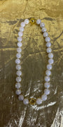 A beaded necklace made of round white pearl-like beads with iridescent blue flashes, accented by two gold-toned spacer beads, displayed on a crinkled gold fabric background.