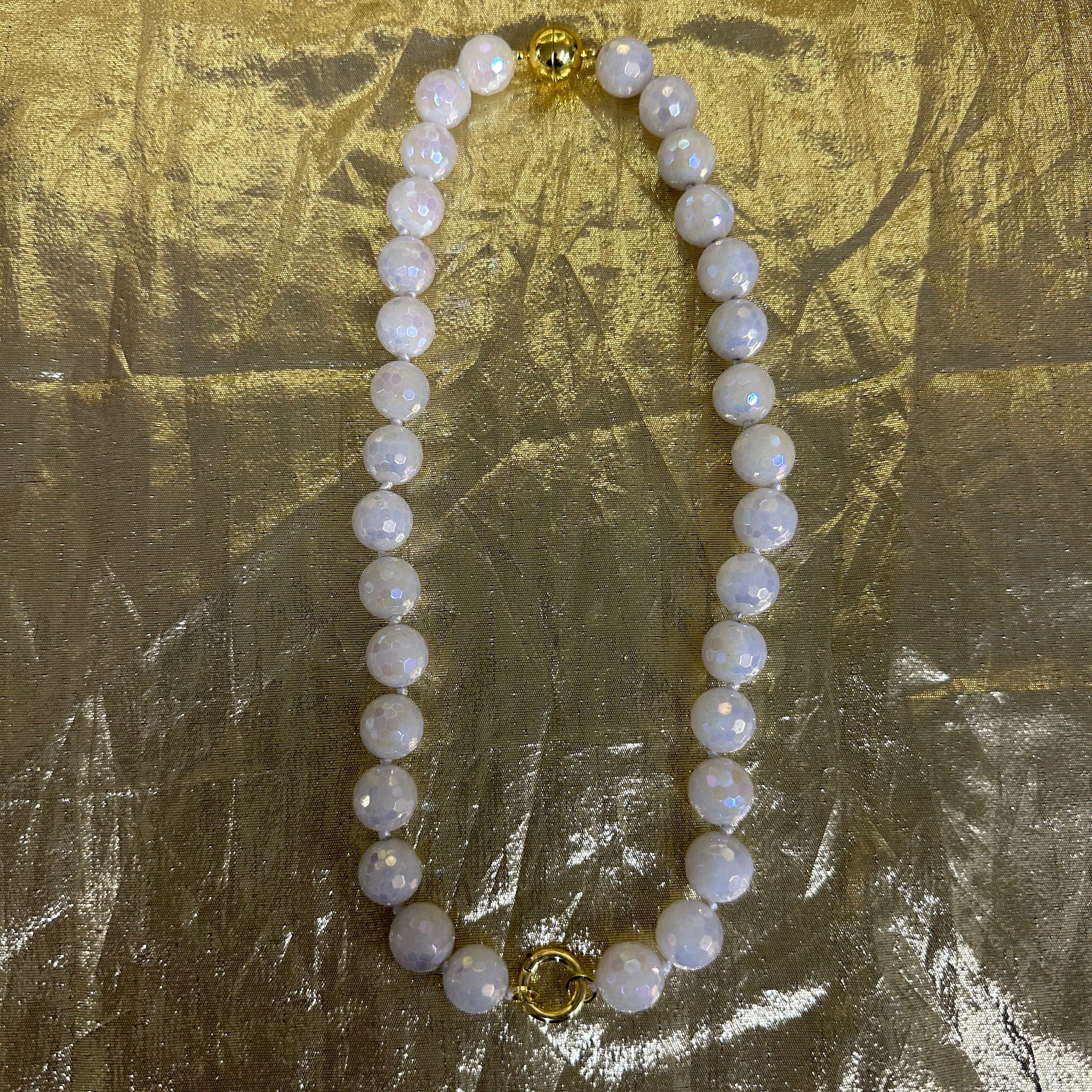 A beaded necklace made of round white pearl-like beads with iridescent blue flashes, accented by two gold-toned spacer beads, displayed on a crinkled gold fabric background.