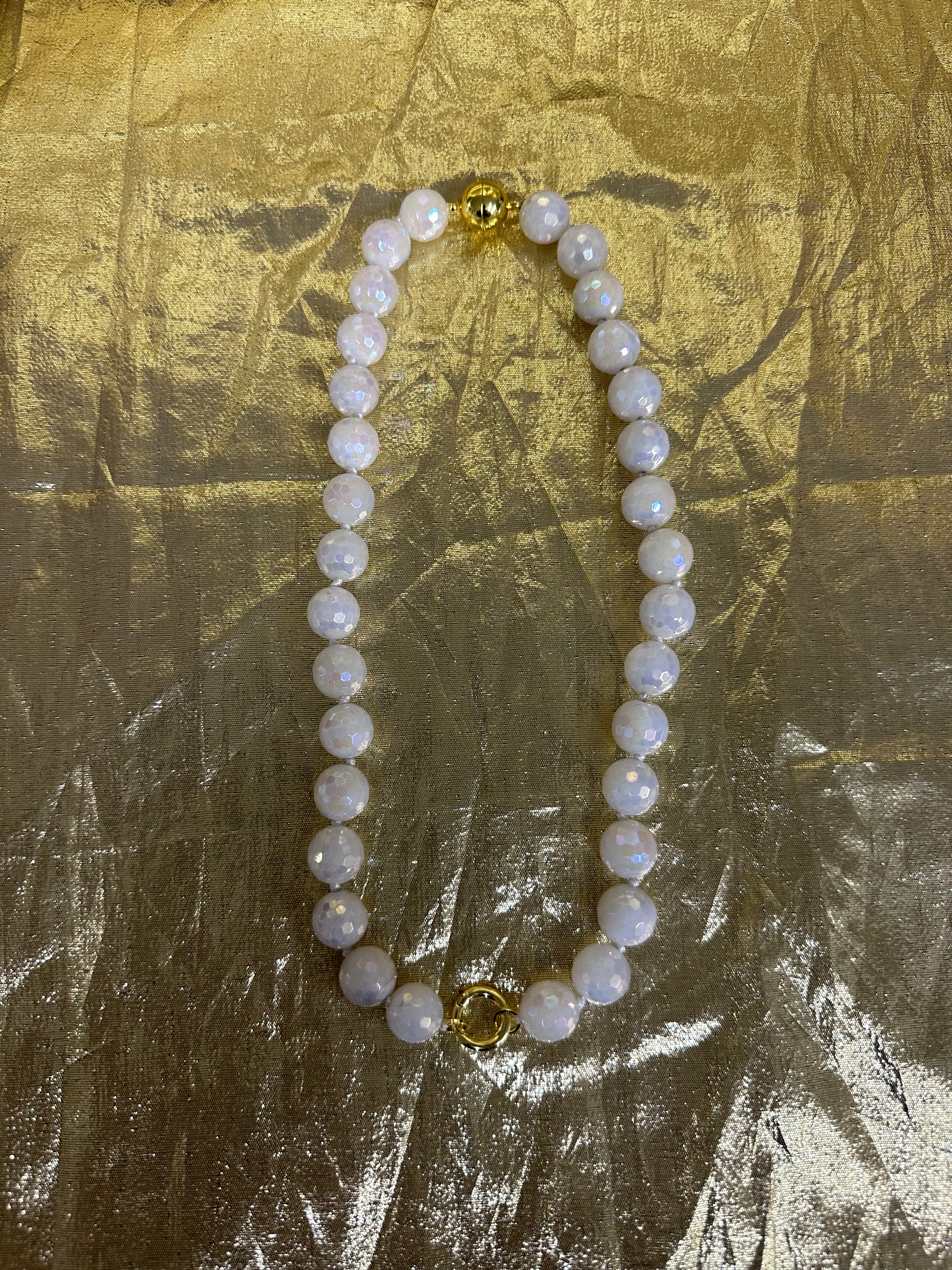 A beaded necklace made of round white pearl-like beads with iridescent blue flashes, accented by two gold-toned spacer beads, displayed on a crinkled gold fabric background.