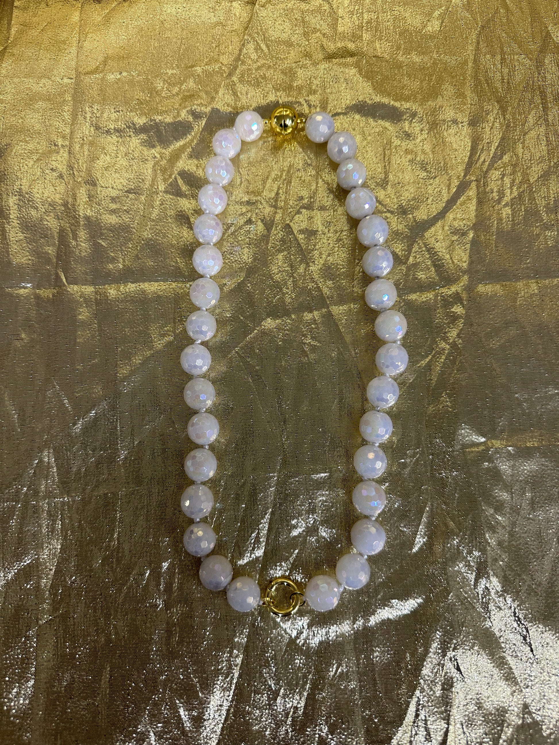 A beaded necklace made of round white pearl-like beads with iridescent blue flashes, accented by two gold-toned spacer beads, displayed on a crinkled gold fabric background.
