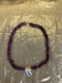 A beaded necklace made of dark purple faceted beads with gold-toned metal accents, including a clasp and a small charm, laid flat on a crinkled gold background. A white product tag is attached near the bottom.
