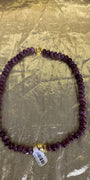 A beaded necklace made of dark purple faceted beads with gold-toned metal accents, including a clasp and a small charm, laid flat on a crinkled gold background. A white product tag is attached near the bottom.