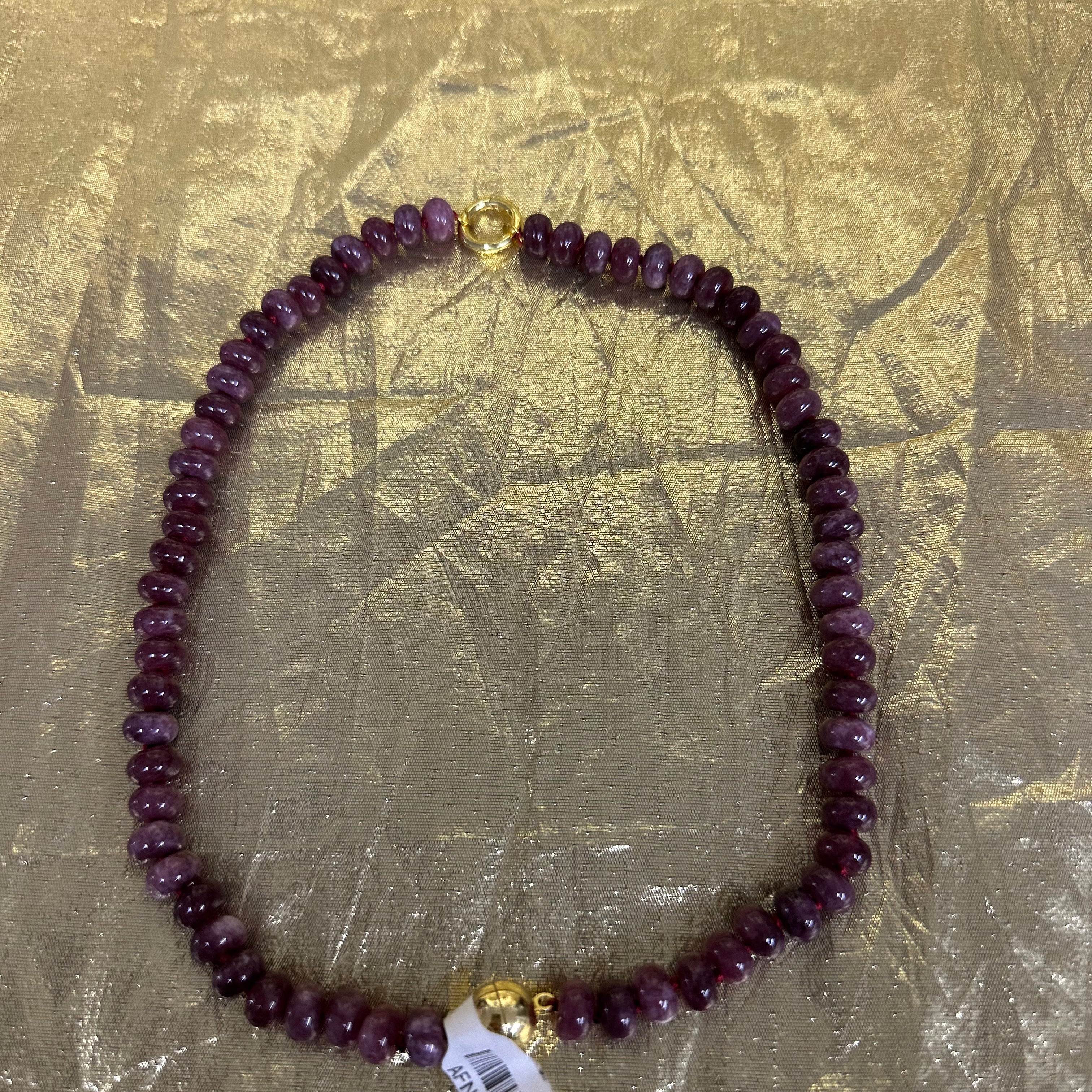 A beaded necklace made of dark purple faceted beads with gold-toned metal accents, including a clasp and a small charm, laid flat on a crinkled gold background. A white product tag is attached near the bottom.
