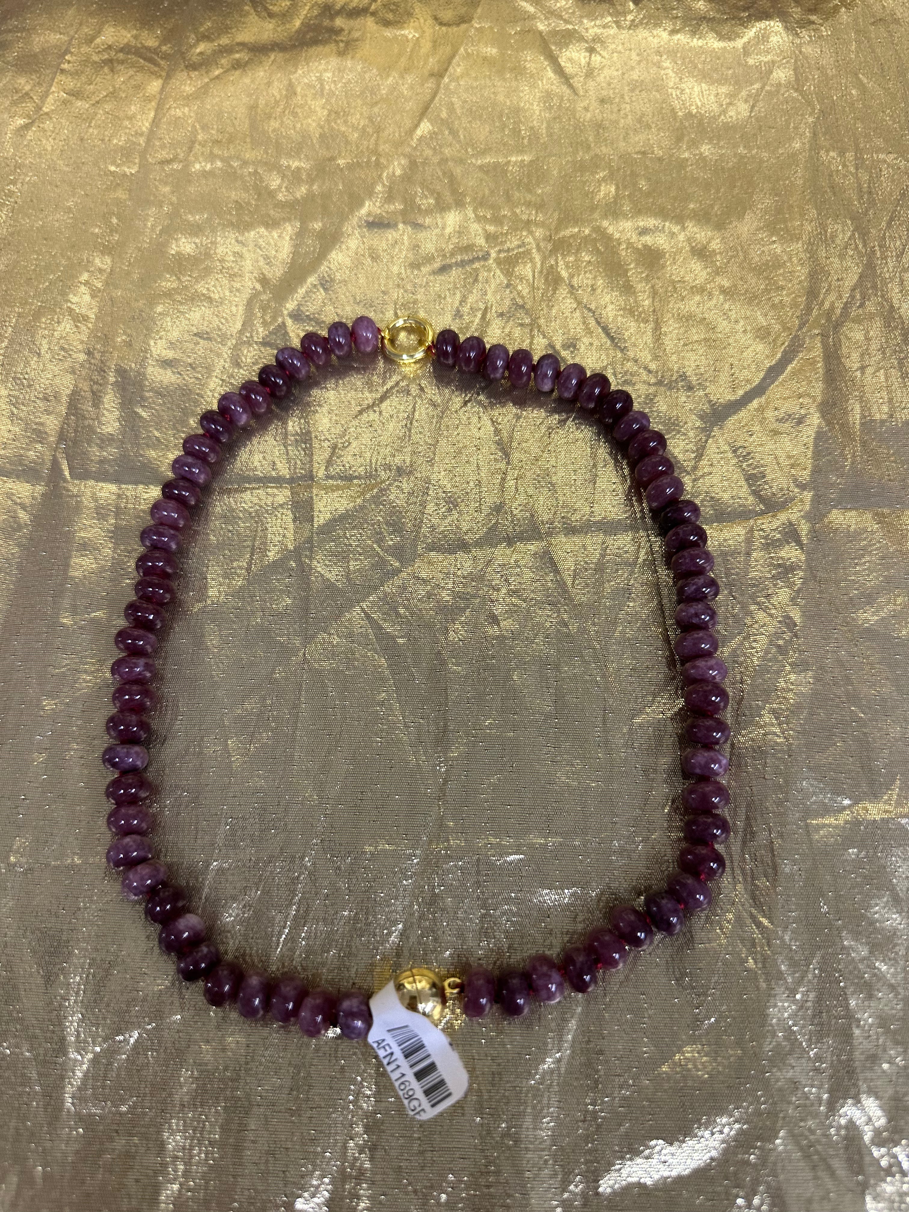 A beaded necklace made of dark purple faceted beads with gold-toned metal accents, including a clasp and a small charm, laid flat on a crinkled gold background. A white product tag is attached near the bottom.