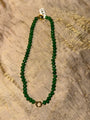 A beaded necklace made of round green faceted beads with a gold-toned spacer bead and a small gold-toned ring clasp, laid flat on a textured beige fabric background.