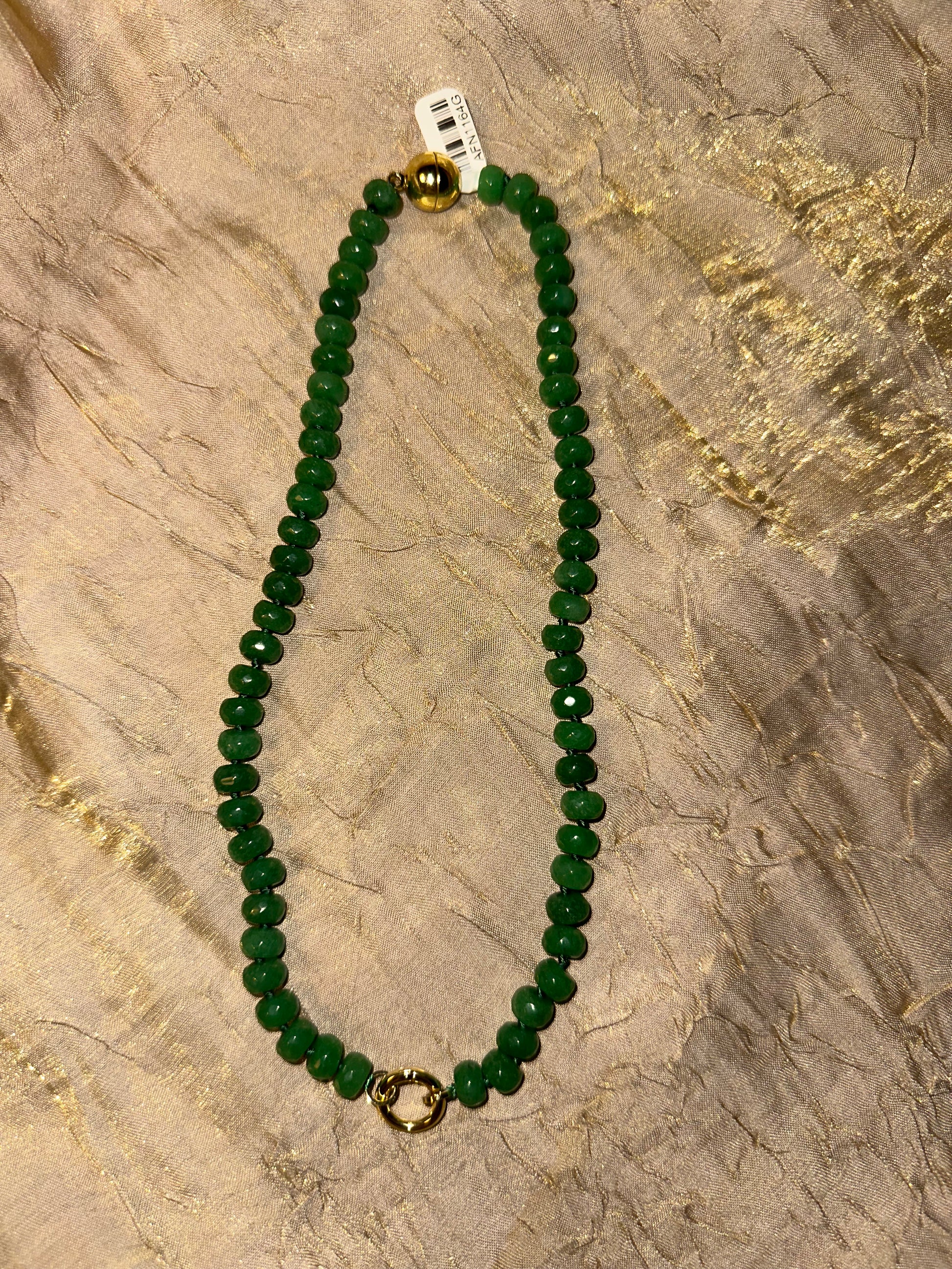 A beaded necklace made of round green faceted beads with a gold-toned spacer bead and a small gold-toned ring clasp, laid flat on a textured beige fabric background.