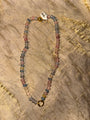 A beaded necklace made of small round multicolored beads in pink, blue, gray, and green tones, with a gold-toned ring clasp and a small white tag attached near the top, laid flat on a textured beige surface.