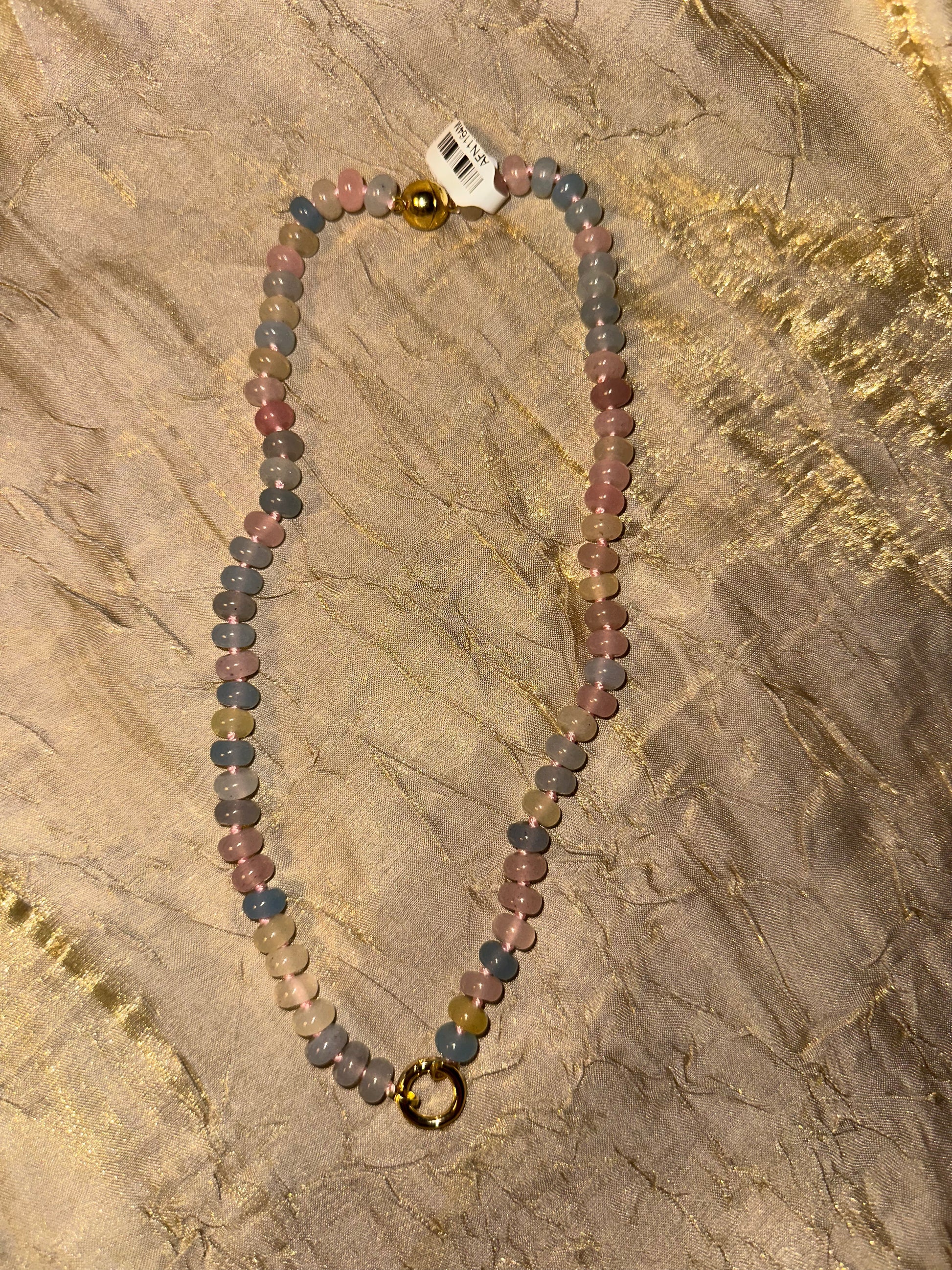 A beaded necklace made of small round multicolored beads in pink, blue, gray, and green tones, with a gold-toned ring clasp and a small white tag attached near the top, laid flat on a textured beige surface.