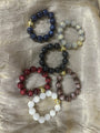 Five beaded stretch bracelets in different colors, including navy blue, light gray, black, red, and brown, with small gold spacer beads, arranged on a textured beige fabric background.