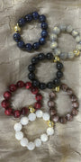 Five beaded stretch bracelets in different colors, including navy blue, light gray, black, red, and brown, with small gold spacer beads, arranged on a textured beige fabric background.