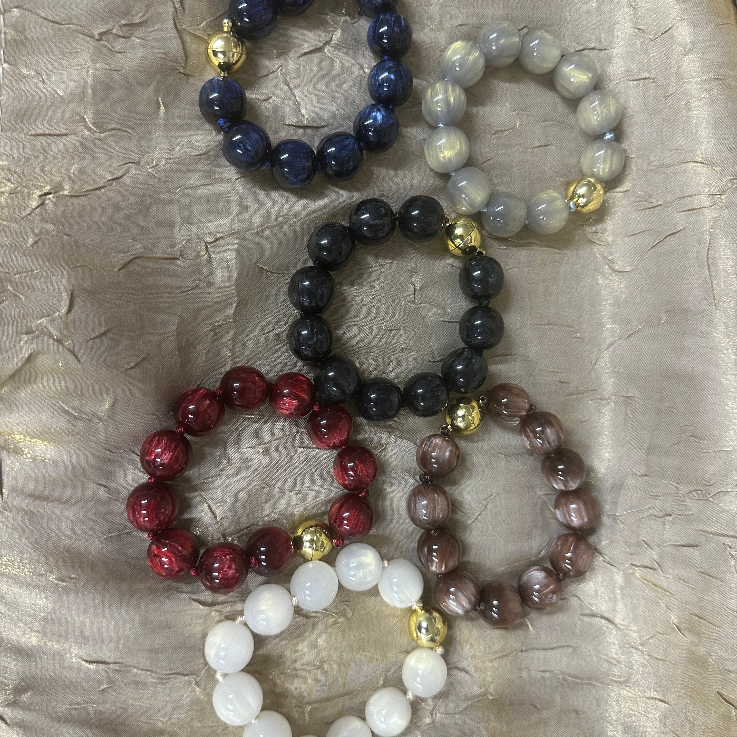 Five beaded stretch bracelets in different colors, including navy blue, light gray, black, red, and brown, with small gold spacer beads, arranged on a textured beige fabric background.