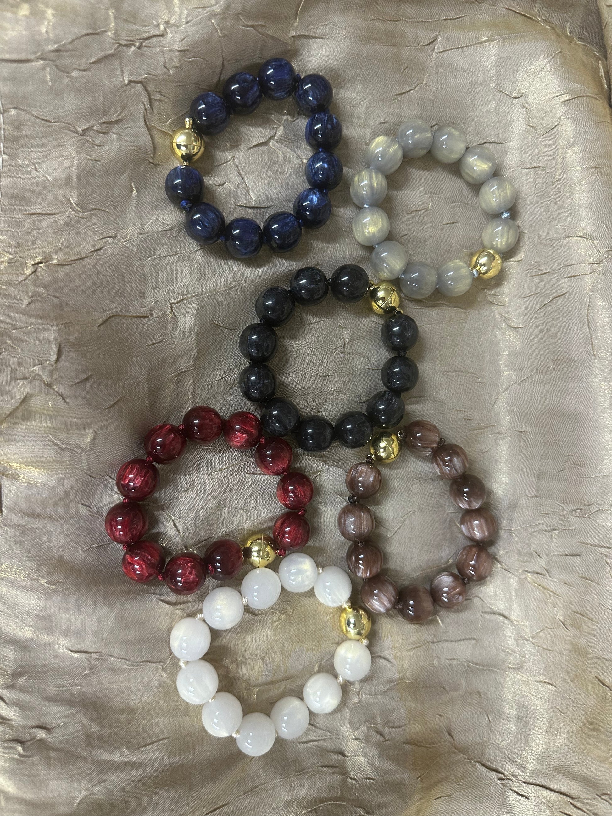 Five beaded stretch bracelets in different colors, including navy blue, light gray, black, red, and brown, with small gold spacer beads, arranged on a textured beige fabric background.
