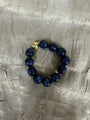 A beaded bracelet made of dark blue lapis lazuli stones with a small gold-toned accent bead, displayed on a textured silver fabric background.