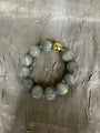 A beaded bracelet made of glossy, marbled gray-green beads with a single metallic gold accent bead, displayed on a textured gray fabric background.
