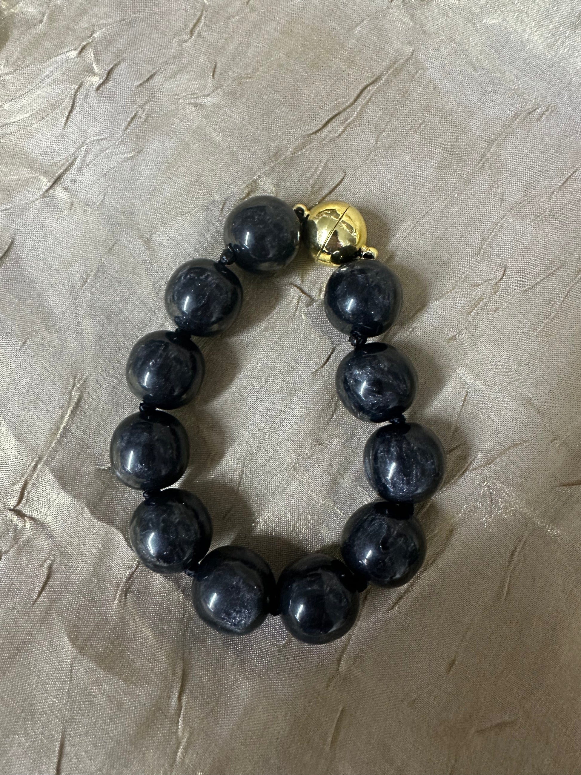 A beaded bracelet made of glossy black round beads with a small gold-toned clasp, photographed on a wrinkled beige fabric background.