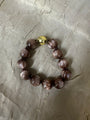 A beaded bracelet made of glossy, marbled brown beads with a single prominent gold-toned bead at the clasp, displayed on a textured beige fabric background.