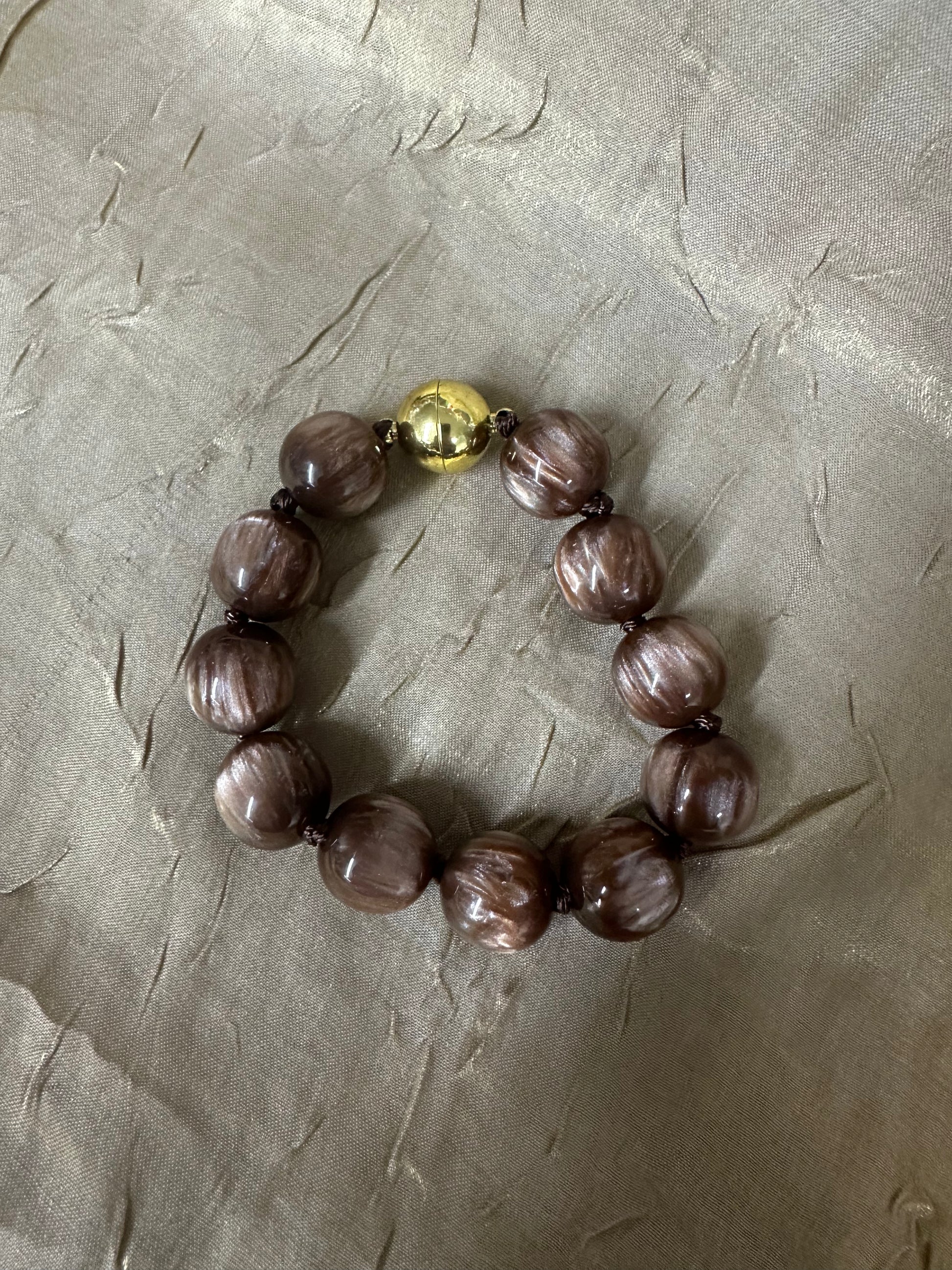 A beaded bracelet made of glossy, marbled brown beads with a single prominent gold-toned bead at the clasp, displayed on a textured beige fabric background.