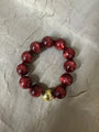 A beaded bracelet made of glossy dark red translucent beads with a small metallic gold accent bead, displayed on a textured light gray surface.