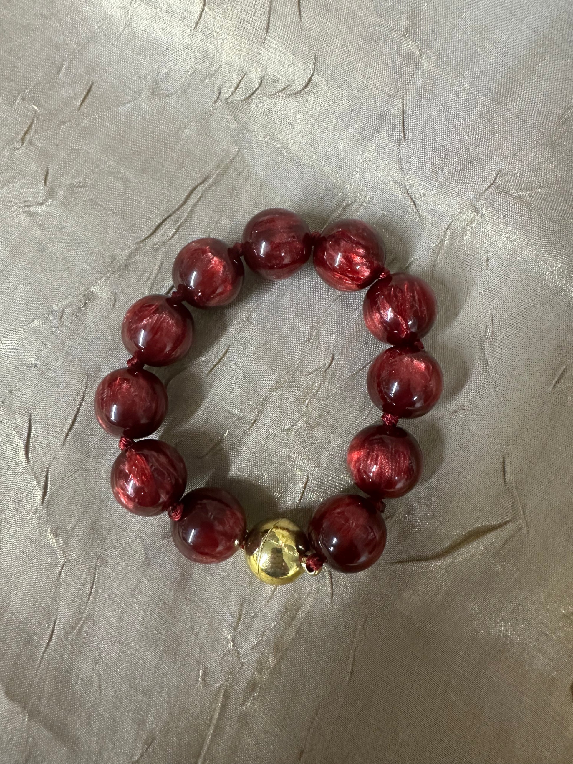 A beaded bracelet made of glossy dark red translucent beads with a small metallic gold accent bead, displayed on a textured light gray surface.