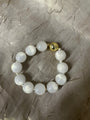 A beaded bracelet made of glossy white round beads with a small gold-toned bell-shaped clasp, displayed on a textured gray fabric background.