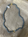 A beaded necklace made of small round blue-gray beads with gold-toned metal accents, including a clasp and a central ring detail, laid on a textured beige fabric background. A small white barcode tag is attached near the top left.