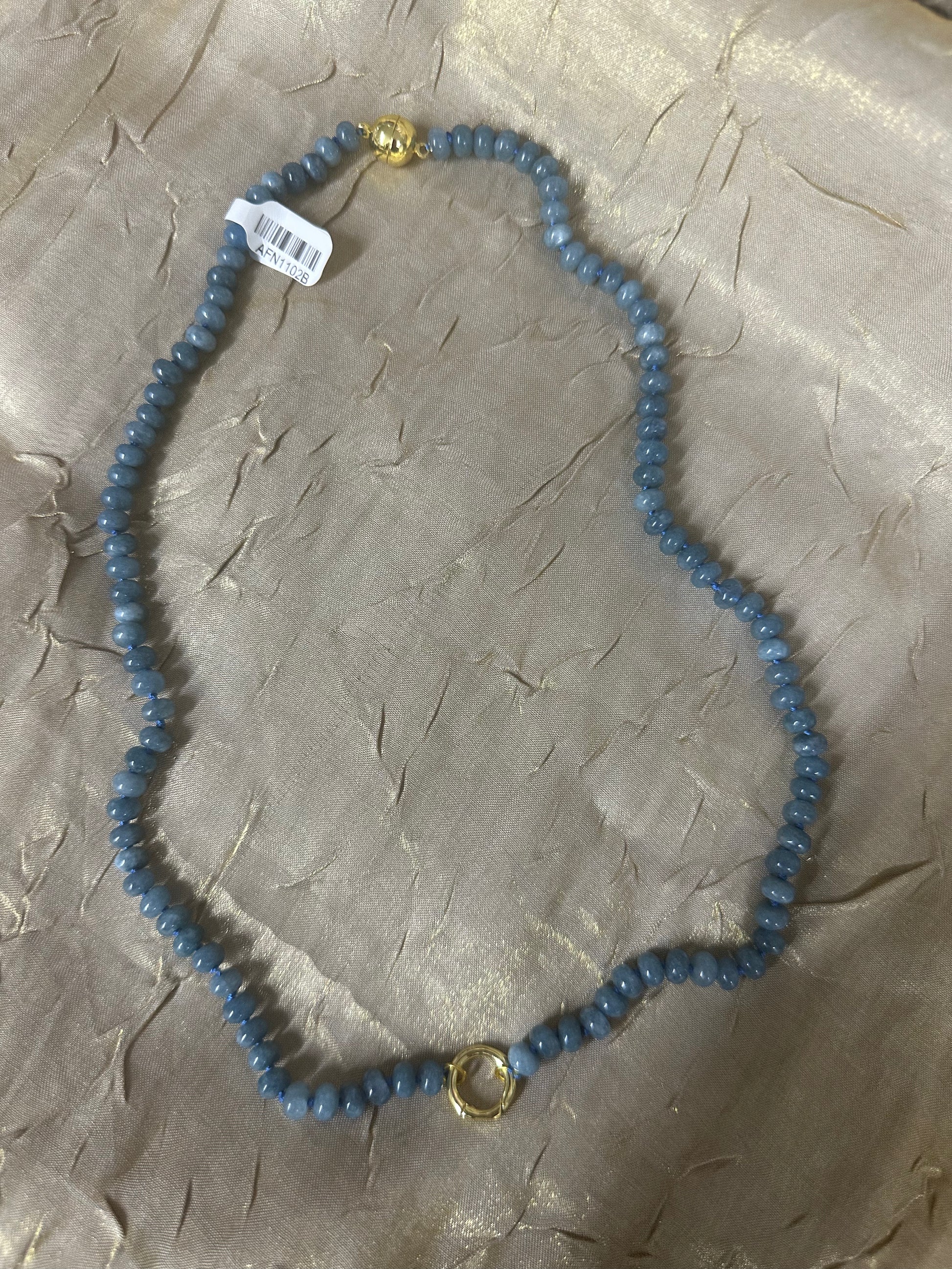 A beaded necklace made of small round blue-gray beads with gold-toned metal accents, including a clasp and a central ring detail, laid on a textured beige fabric background. A small white barcode tag is attached near the top left.