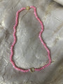 A pink beaded necklace with small faceted beads and gold-toned metal accents, laid flat on a textured light gray fabric background.