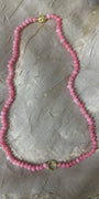 A pink beaded necklace with small faceted beads and gold-toned metal accents, laid flat on a textured light gray fabric background.