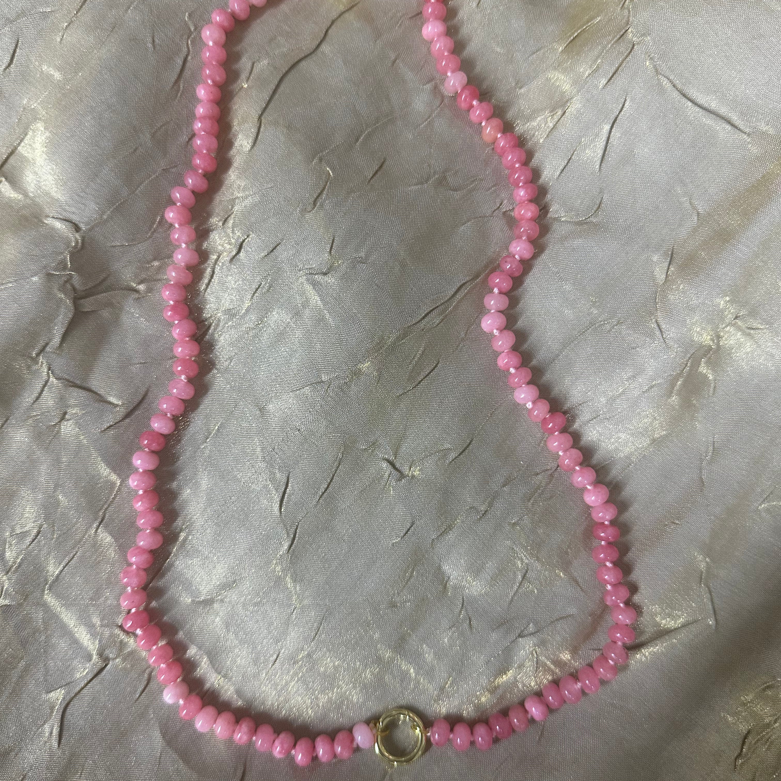 A pink beaded necklace with small faceted beads and gold-toned metal accents, laid flat on a textured light gray fabric background.