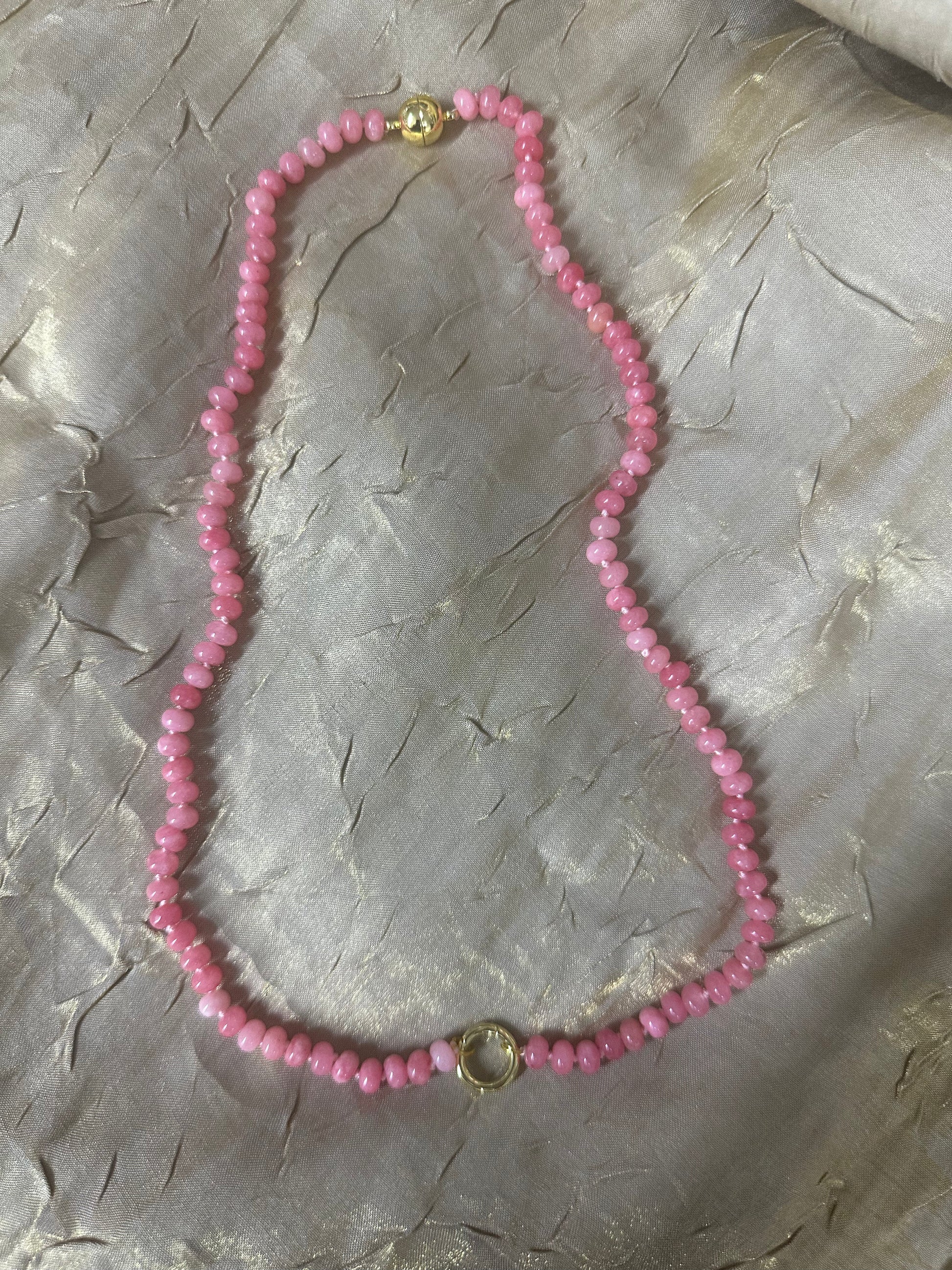 A pink beaded necklace with small faceted beads and gold-toned metal accents, laid flat on a textured light gray fabric background.