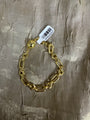 A gold chain bracelet with large paperclip-style links and a lobster clasp, shown with a white barcode tag on a textured metallic taupe fabric background.