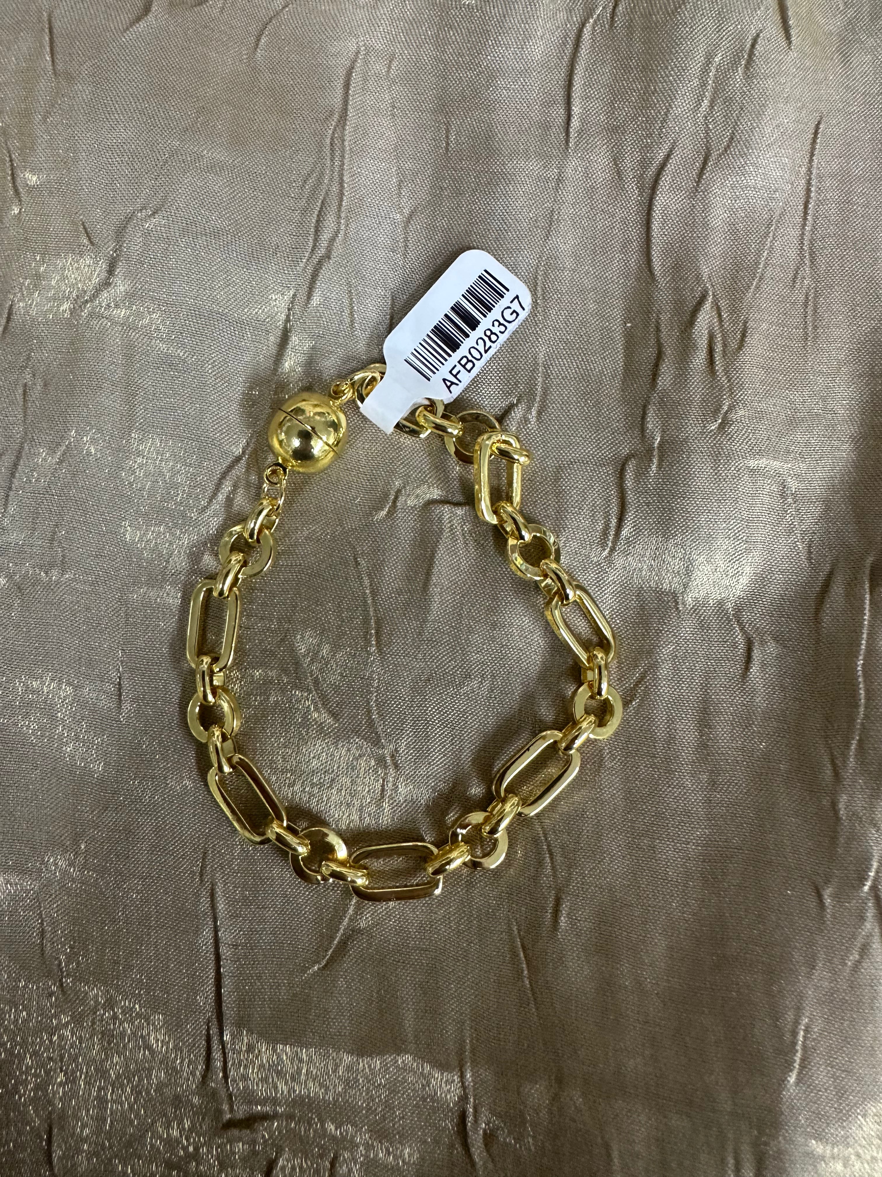 A gold chain bracelet with large paperclip-style links and a lobster clasp, shown with a white barcode tag on a textured metallic taupe fabric background.