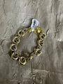A gold and silver chain bracelet with large interlocking gold rings and a small gold ball charm, shown on a textured metallic taupe fabric background with a white product tag attached.