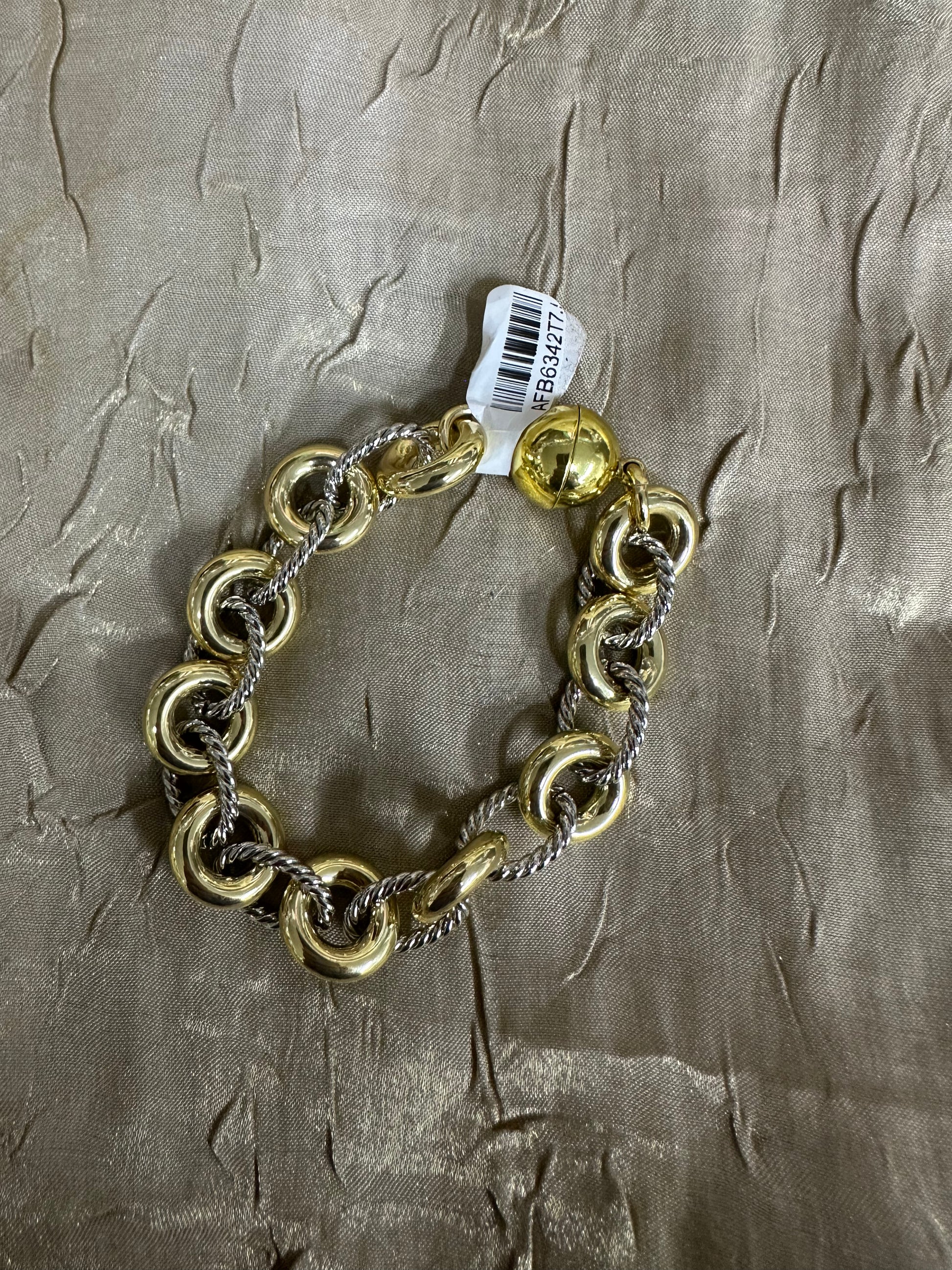 A gold and silver chain bracelet with large interlocking gold rings and a small gold ball charm, shown on a textured metallic taupe fabric background with a white product tag attached.