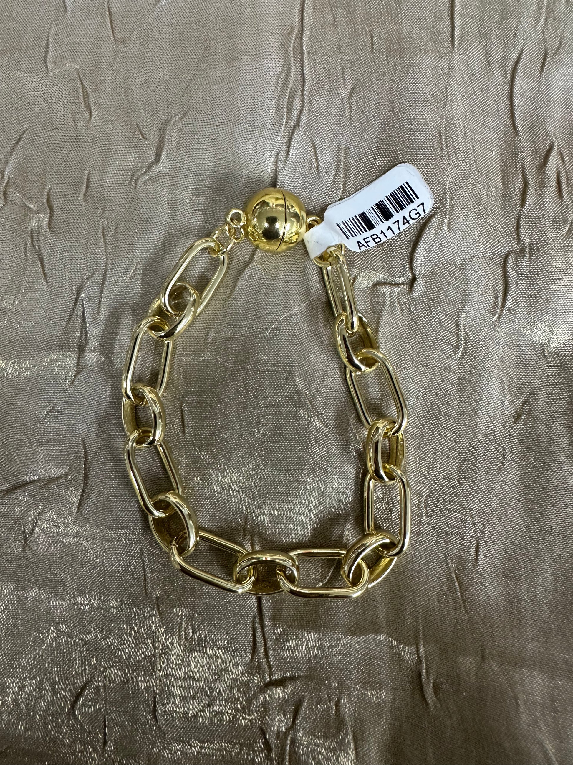 A gold-toned chunky chain link bracelet with a spherical clasp and a white barcode tag, displayed on a textured metallic gold fabric background.