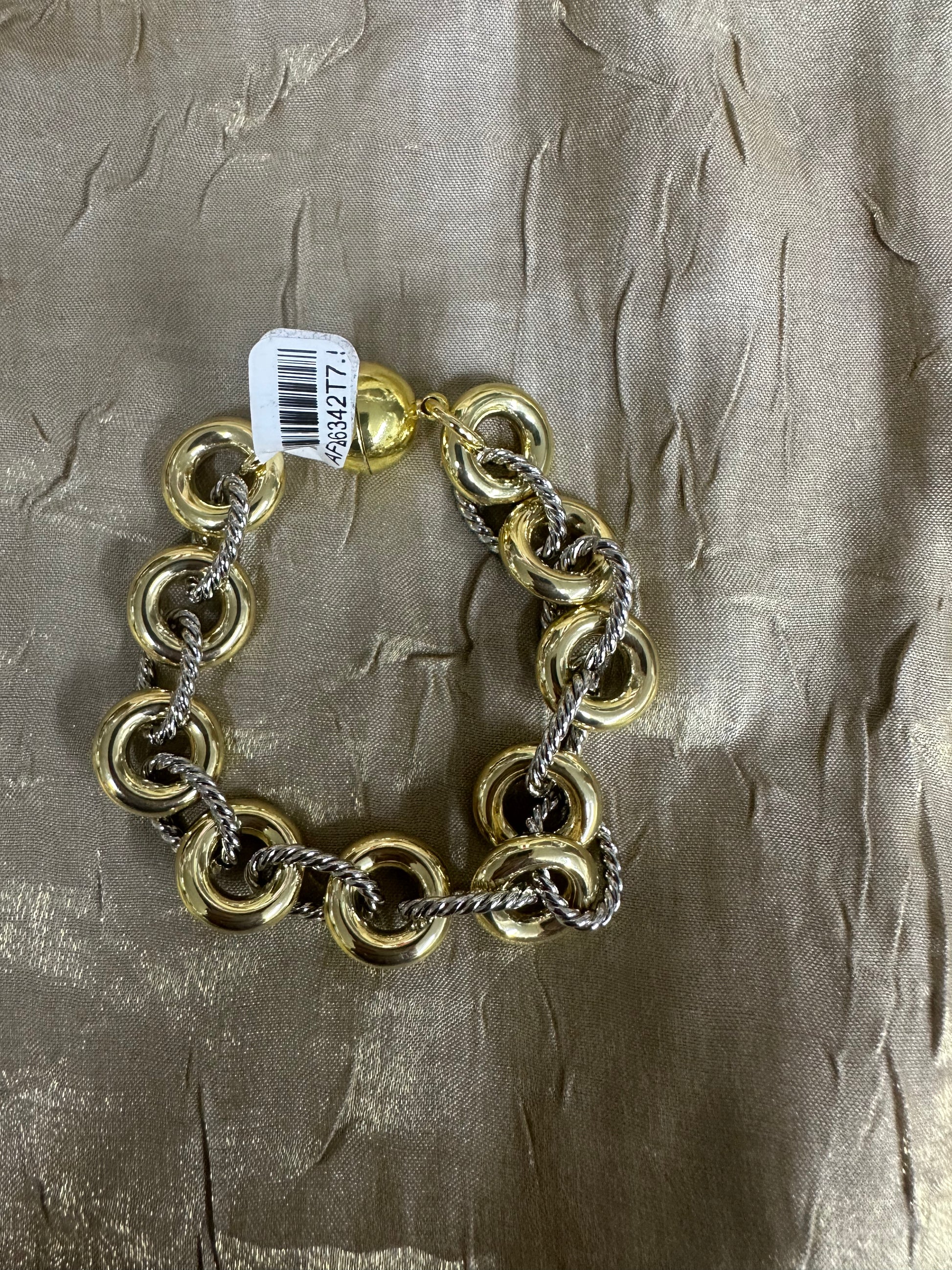 A gold and silver-tone chain link bracelet with large interlocking rings, shown on a textured metallic gold fabric background with a white barcode tag attached.