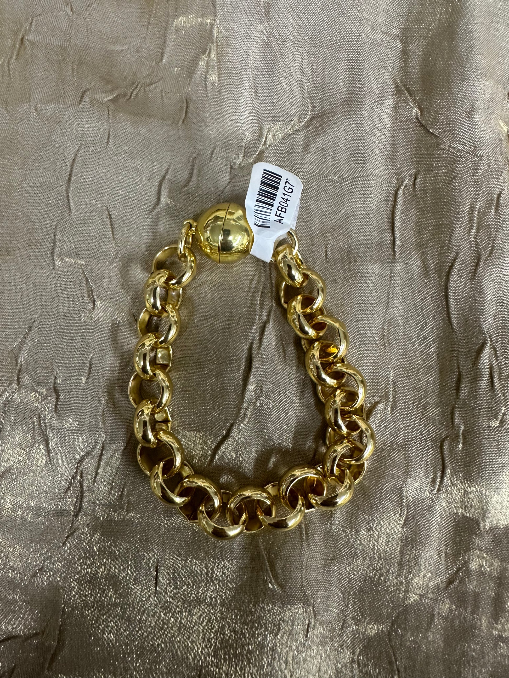 A chunky gold chain bracelet with a matching gold ball charm, resting on a textured metallic gold fabric background with a white barcode tag attached.