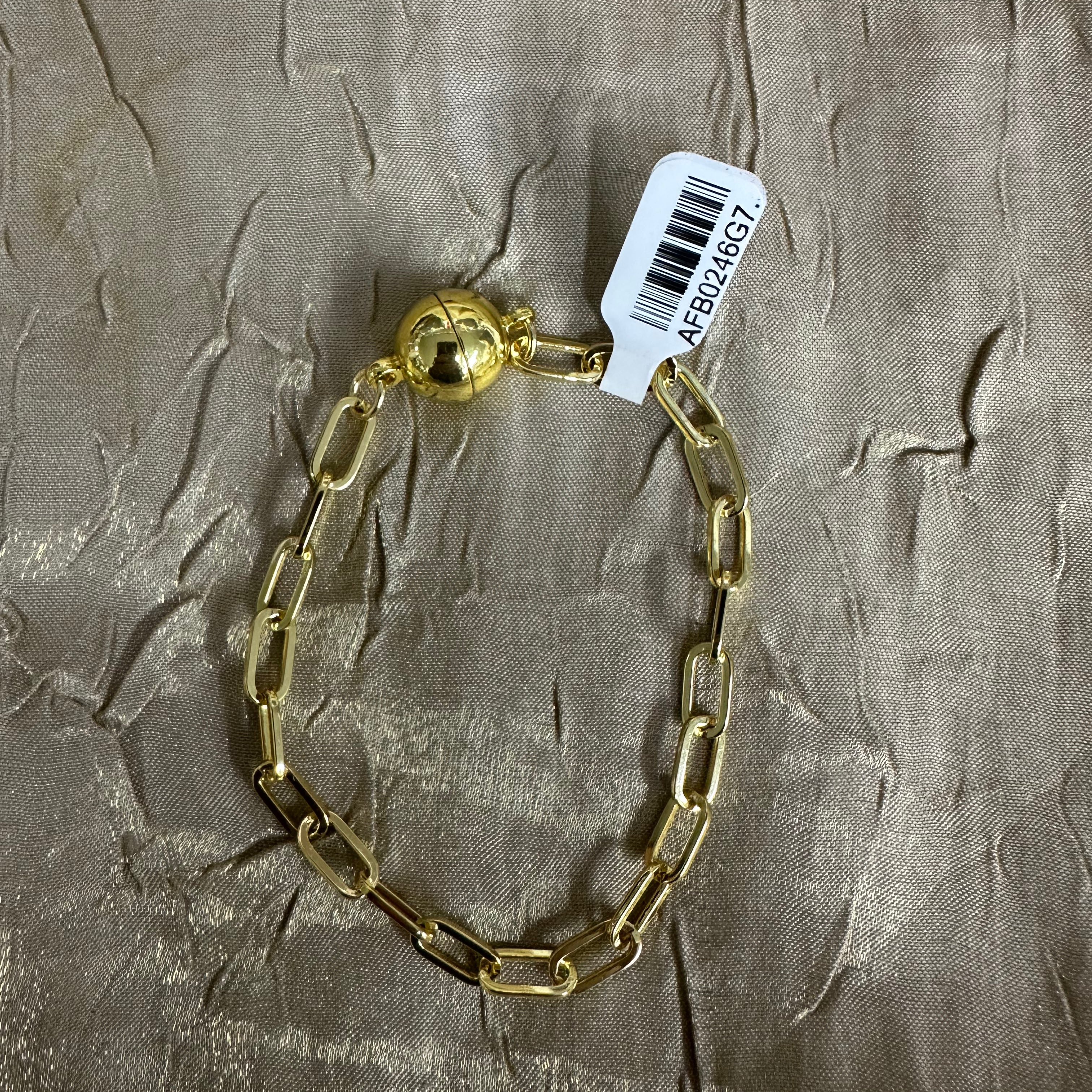 A gold chain bracelet with a polished ball clasp, laid flat on a textured beige fabric background with a white product tag attached.
