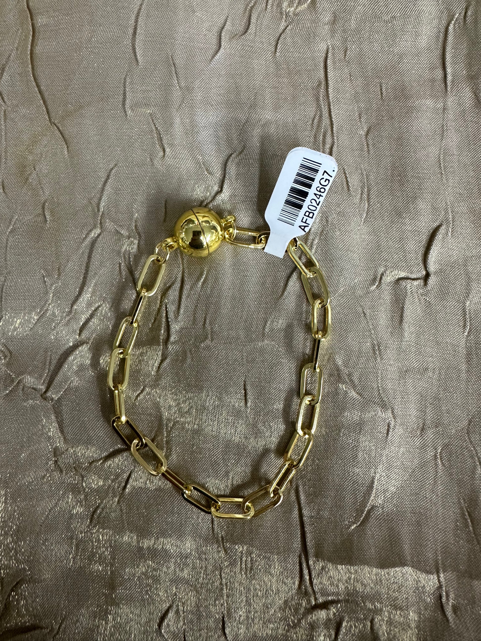 A gold chain bracelet with a polished ball clasp, laid flat on a textured beige fabric background with a white product tag attached.