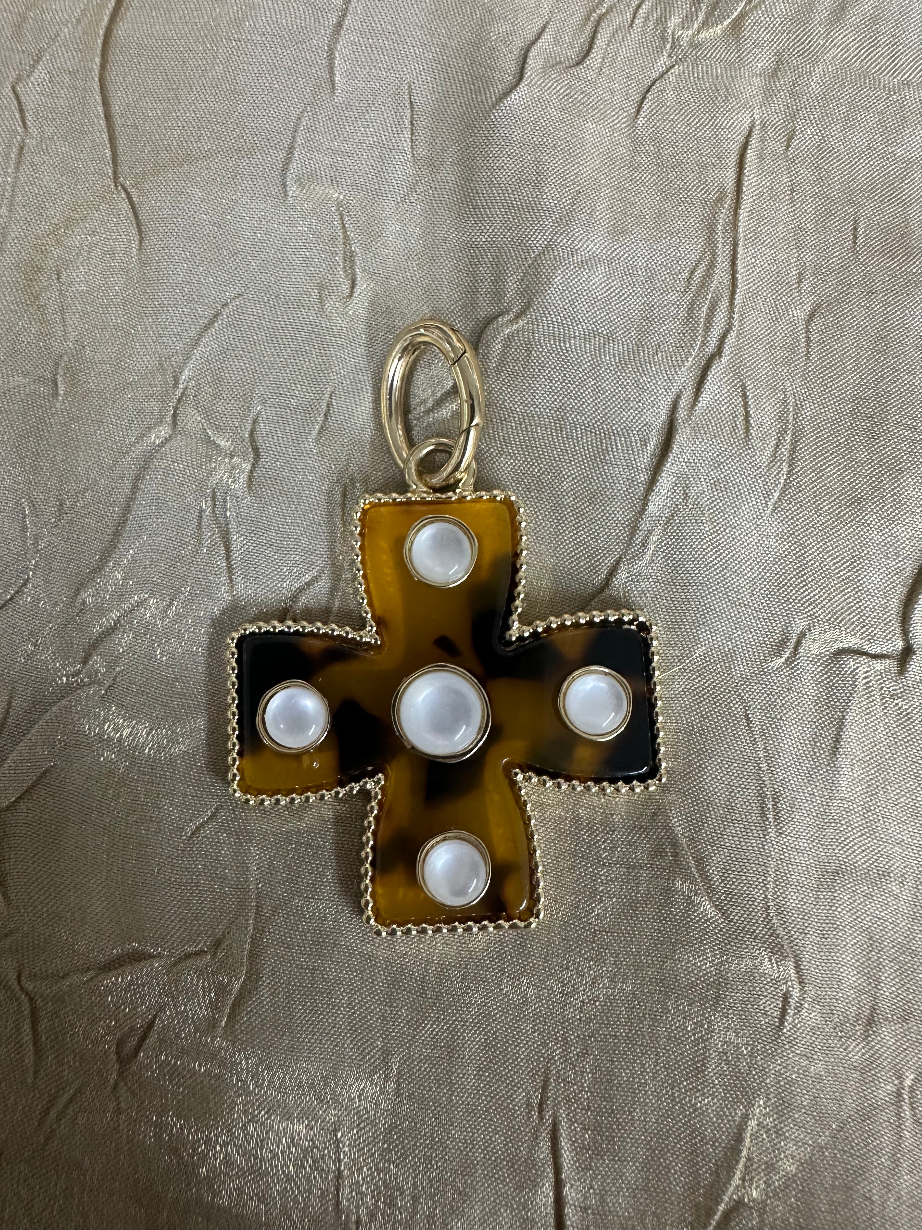 A vintage-style cross pendant featuring a tortoiseshell pattern in amber-brown and black, accented with five white faux pearls and a gold-tone bail, displayed on a textured beige fabric background.