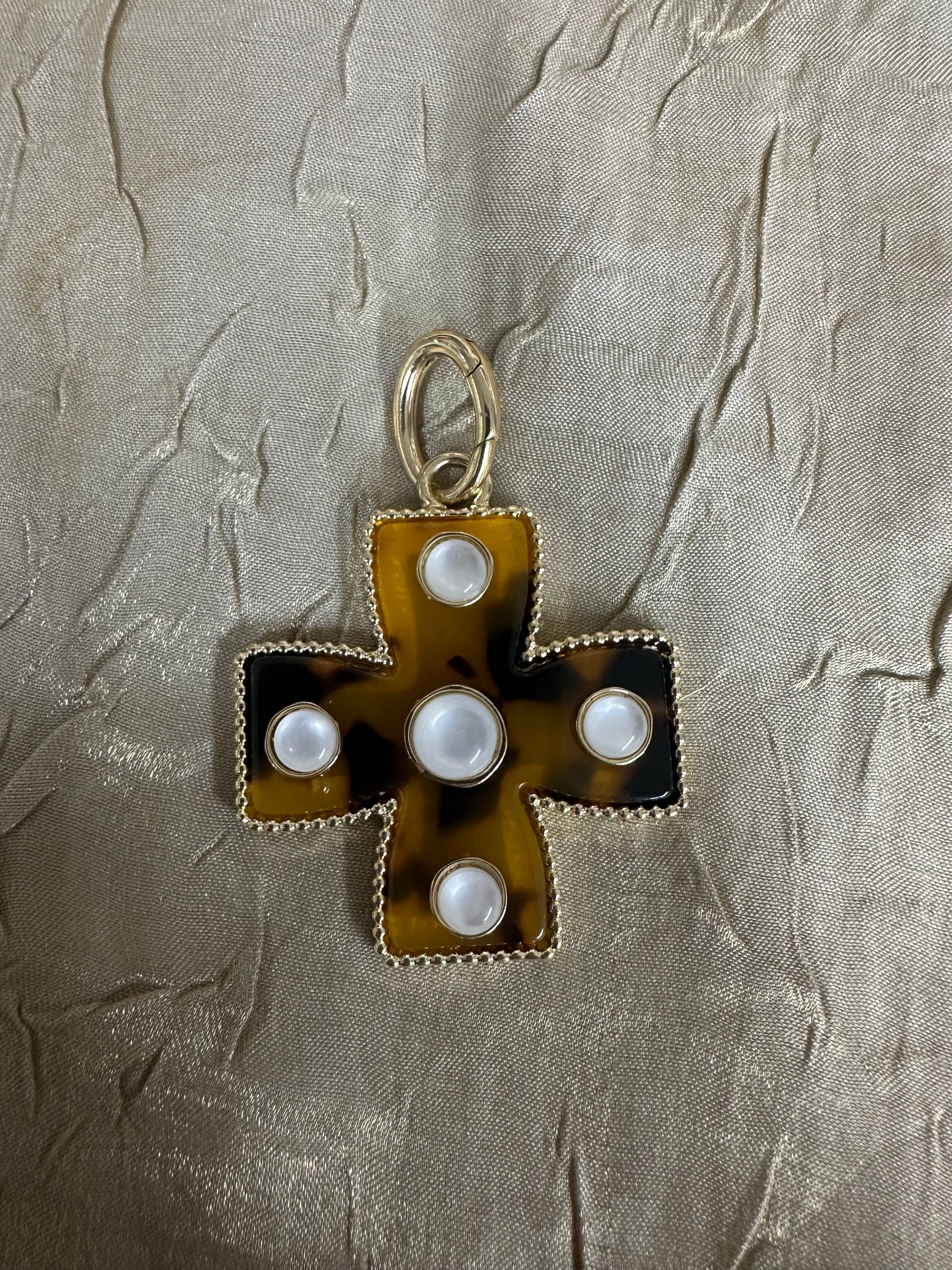 A vintage-style cross pendant featuring a tortoiseshell pattern in amber-brown and black, accented with five white faux pearls and a gold-tone bail, displayed on a textured beige fabric background.
