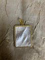 A rectangular pendant featuring a polished mother-of-pearl inlay with iridescent gray and white tones, set in a gold-tone beaded frame with a gold bail, displayed on a textured gold fabric background.