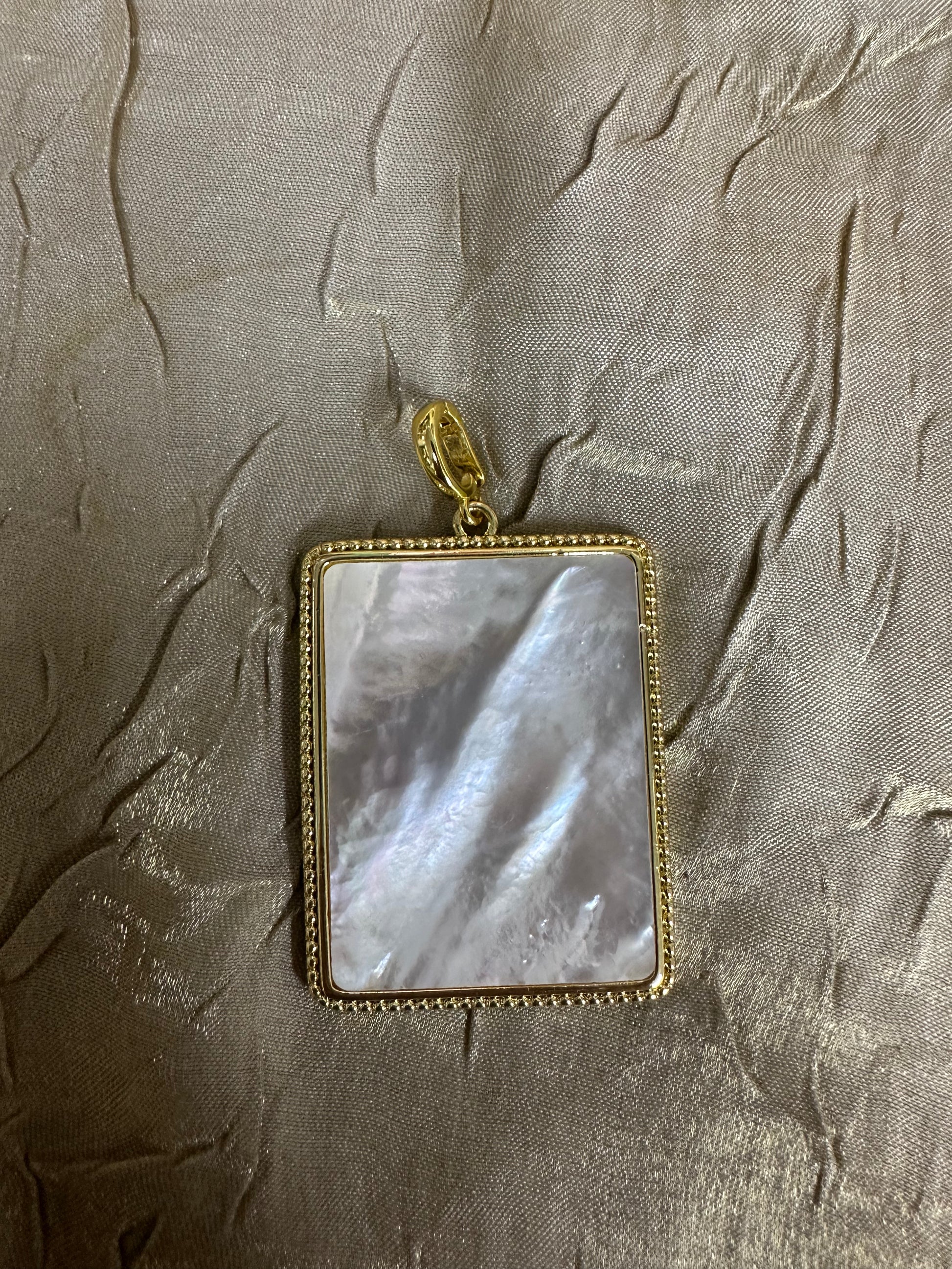 A rectangular pendant featuring a polished mother-of-pearl inlay with iridescent gray and white tones, set in a gold-tone beaded frame with a gold bail, displayed on a textured gold fabric background.