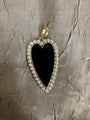 A heart-shaped pendant featuring a glossy black center, bordered by small white faux pearls and a gold-tone metal frame, resting on a textured beige fabric background.